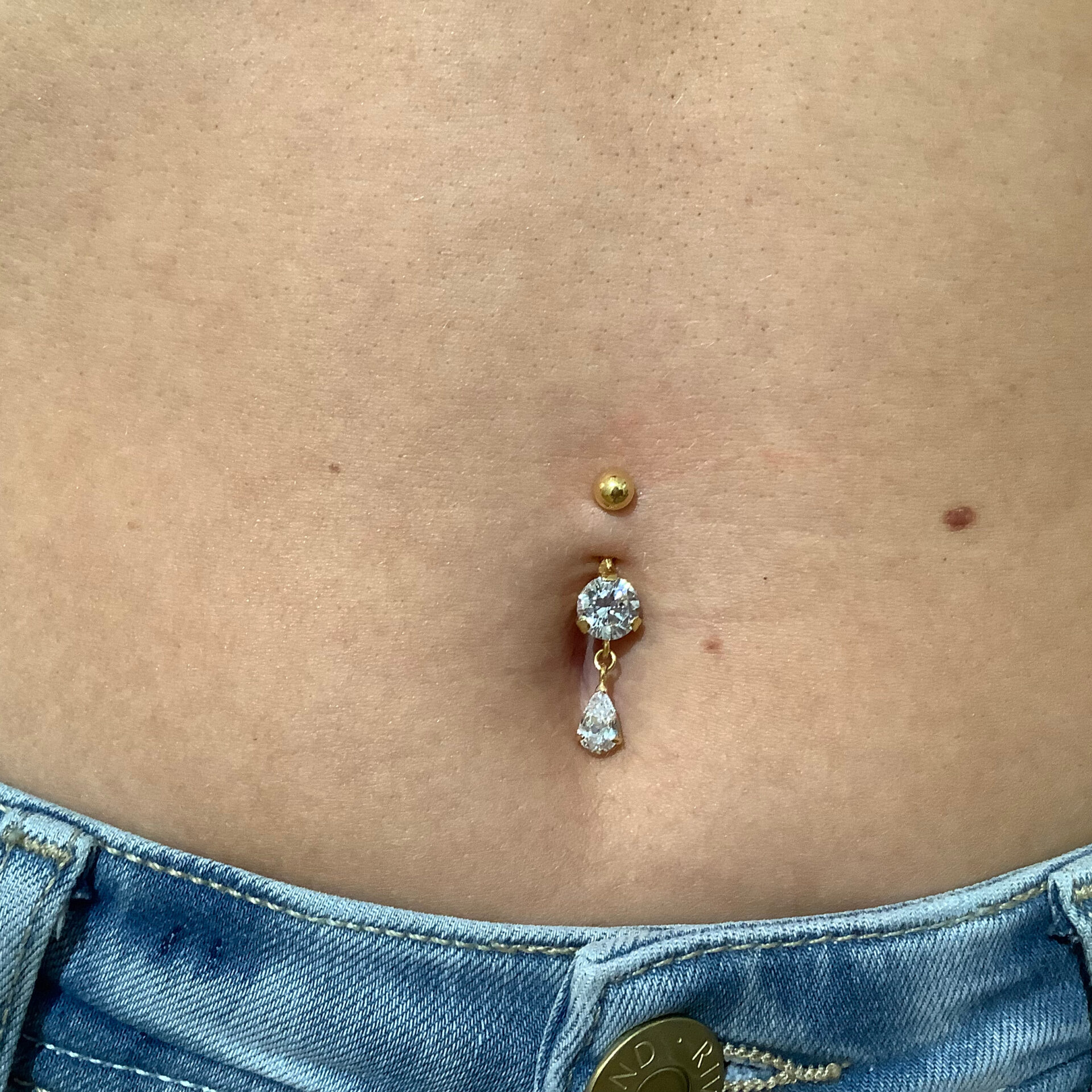 Schmuckstyling von Miri. Großer Piercingschmuck in Gold mit großen tropfenformenen Kristallen in Bauchnabelpiercing. Dieser stilvolle goldene Piercingschmuck mit glitzernden tropfenförmigen Steinchen wurde in unserem Studio in ein vorhandenes Bauchnabelpiercing eingesetzt.